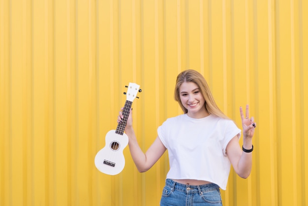 Jolie blonde modèle avec un ukulélé blanc dans ses mains pose sur un mur jaune.