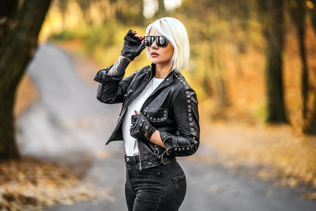 Jolie blonde de style motard femmes en veste de cuir noir avec des lunettes de soleil debout à l'extérieur dans la forêt