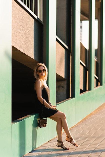 Une jolie blonde vêtue d'une robe d'été avec un sac à main est fatiguée de se promener dans la ville et de s'asseoir pour se reposer