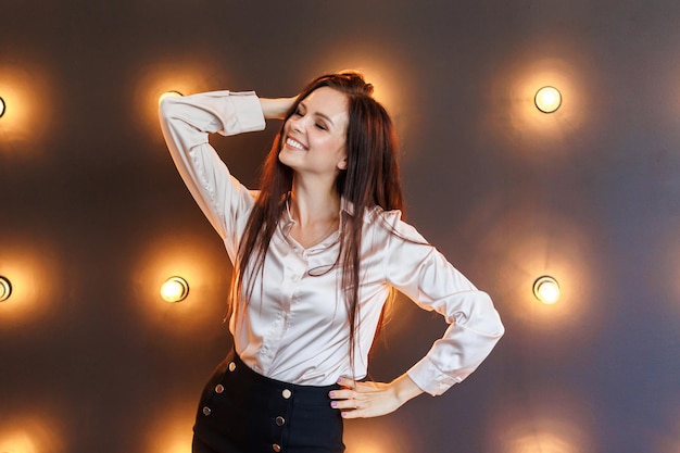 Jolie brune en chemisier et jupe courte se tient en studio avec un mur de briques et des lampes et garde ses mains sur sa tête