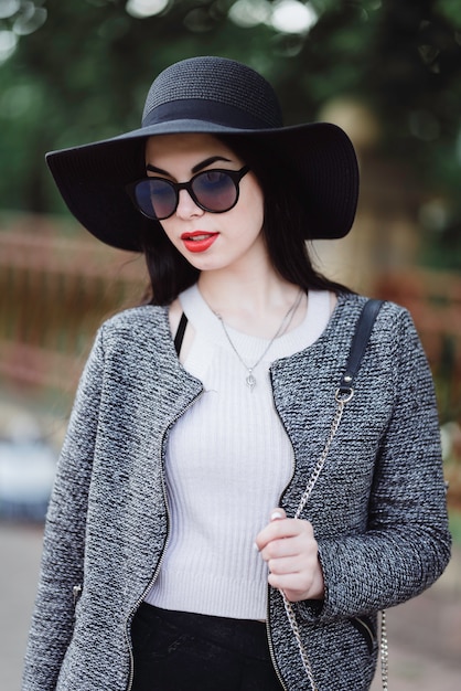 Jolie brune avec des lèvres rouges au chapeau et des lunettes de soleil posant dans la ville