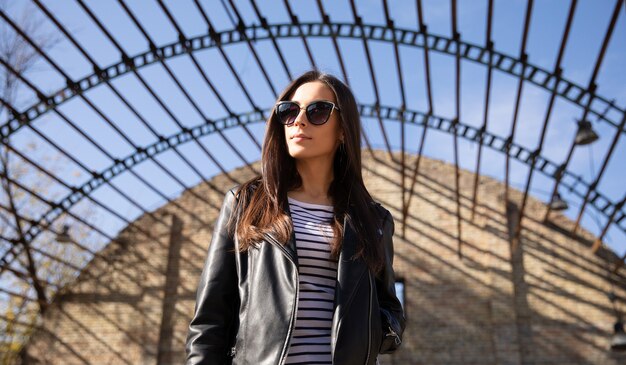 Jolie brune pose en semi-profil sur fond urbain, portant des lunettes de soleil, une veste en cuir et une chemise rayée