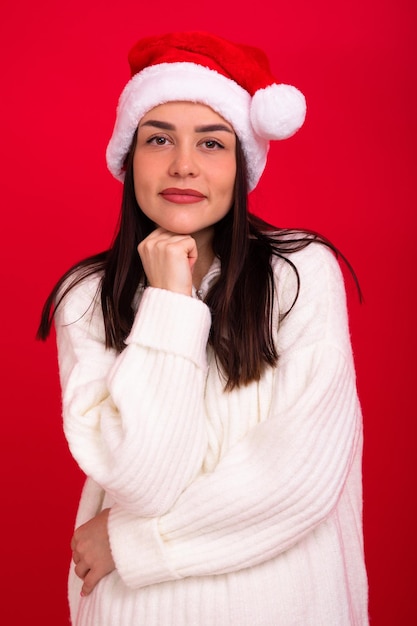 Jolie brune en pull blanc et chapeau de père Noël sur fond rouge Le modèle regarde la caméra