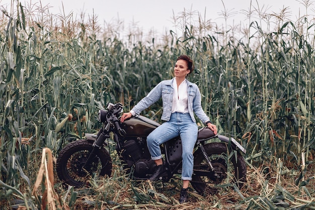 Jolie dame adulte et séduisante portant des vêtements en jean et assise sur une grande moto rétro noire sur fond d'herbe et de verdure. Concert de tourisme