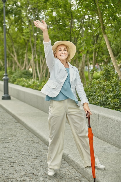 Jolie dame âgée blanche d'environ 62 ans voyage en se tenant debout sur le trottoir dans un parc public.