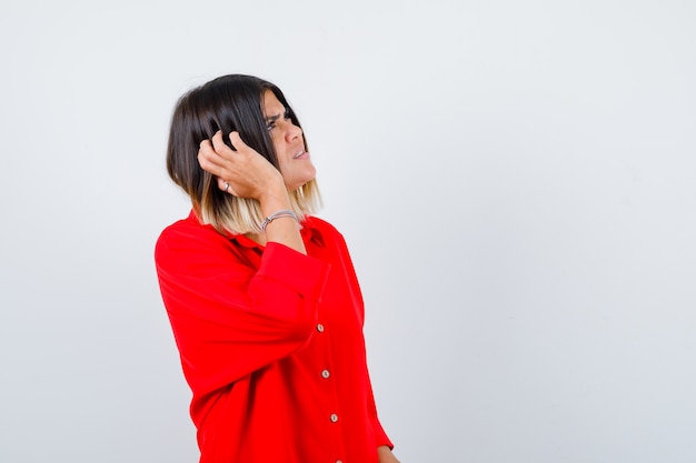 Photo jolie dame en chemisier rouge se grattant la tête, levant les yeux et l'air oublieux, vue de face.