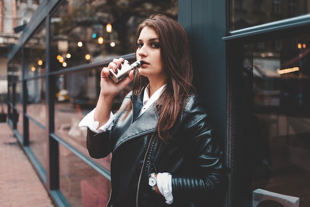 Jolie dame dans une veste en cuir noir fume une cigarette électronique près des fenêtres en verre