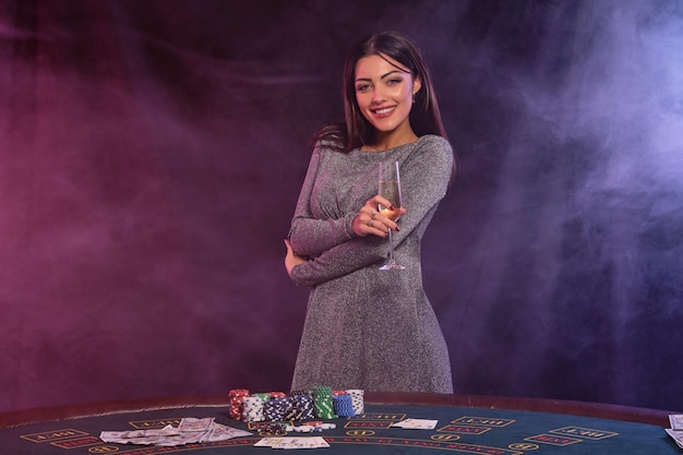 Jolie dame en robe grise jouant au poker au casino tenant un verre de champagne Posant à table avec des piles de jetons cartes d'argent dessus Fumée noire rétro-éclairage coloré Gros plan