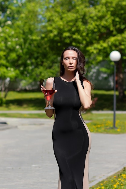 Jolie dame en robe noire élégante touchant le cou et dégustant du vin rouge le jour d'été ensoleillé