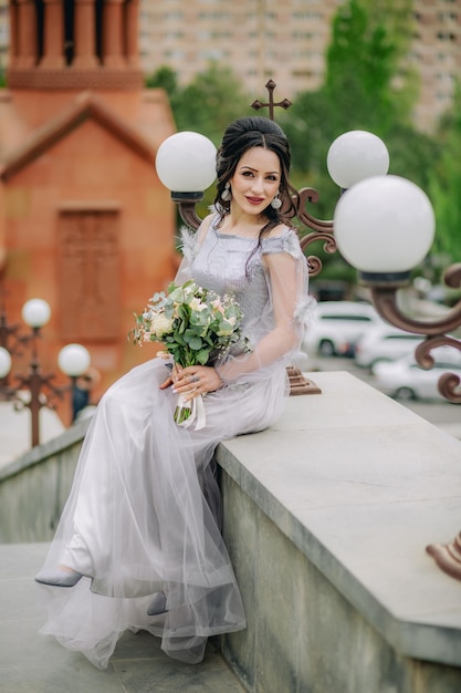Photo jolie demoiselle d'honneur souriante assise sur une pierre devant l'église tenant un bouquet de mariée