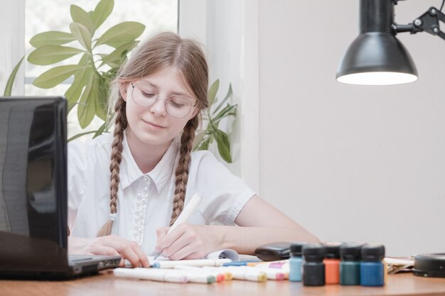 Jolie écolière étudiant ses devoirs pendant sa leçon en ligne à la maison, distance sociale pendant la quarantaine, auto-isolement, concept d'éducation en ligne, élève à domicile. Enseignement à distance en ligne