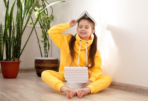 Photo jolie écolière sourit et tient un livre sur la tête posant devant la caméra dans sa chambre concept de l'éducation l'enfant apprend à lire et à écrire concept d'éducation devoirs mode de vie