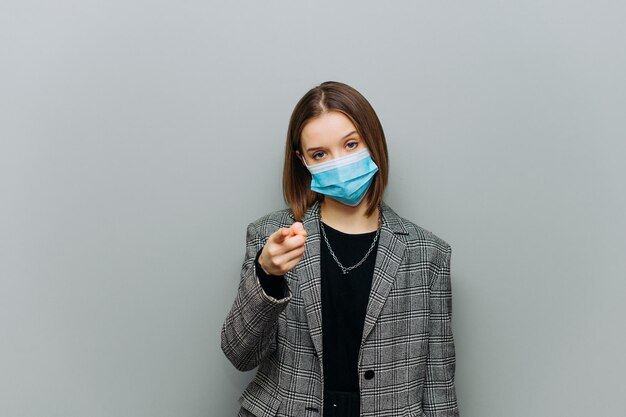 Jolie employée de bureau en costume et masque médical sur le visage montrant le doigt à la caméra