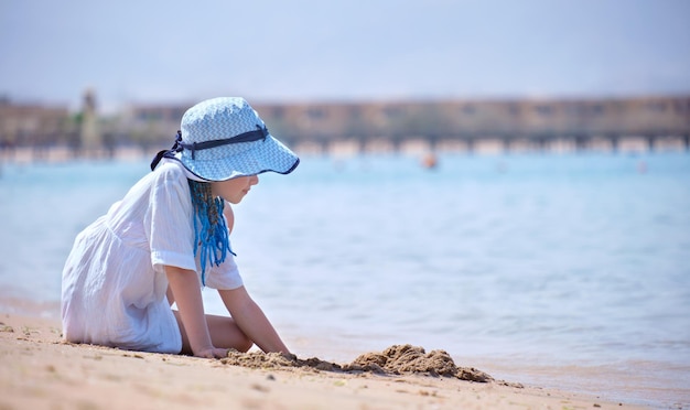 Une jolie enfant dans un grand chapeau et une robe blanche jouant avec du sable mouillé sur le fond du ciel bleu et de l'eau claire de l'océan lagon