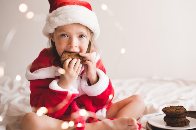 Jolie enfant fille porter un chapeau de père noël rouge et un peignoir s'asseoir dans son lit avec un décor de Noël