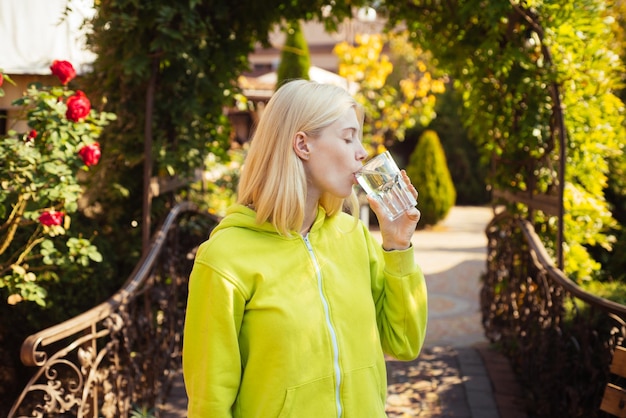 Jolie étudiante aux cheveux blonds habillée avec désinvolture en train de boire un verre d'eau en étant heureuse dehors...