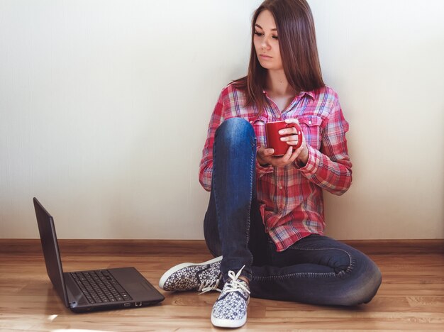 Une jolie étudiante est assise sur le sol avec un ordinateur portable et une tasse de café. Enseignement à la maison concept