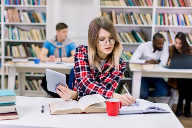 Jolie étudiante de race blanche ciblée assis dans un espace de coworking étudier avec un livre et une tablette, prendre des notes et se préparer pour un test ou un examen en bibliothèque