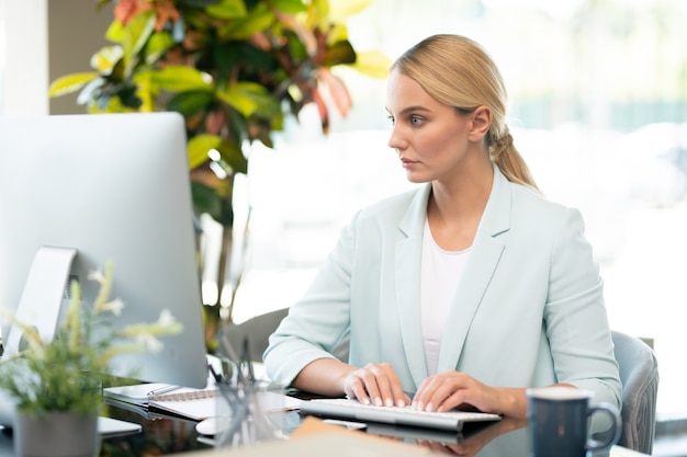 Jolie étudiante se concentrant sur la lecture d'informations en ligne sur l'écran de l'ordinateur tout en tapant par bureau