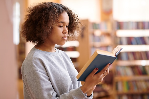 Jolie étudiante en tenue décontractée lisant un livre intéressant tout en visitant la bibliothèque du collège