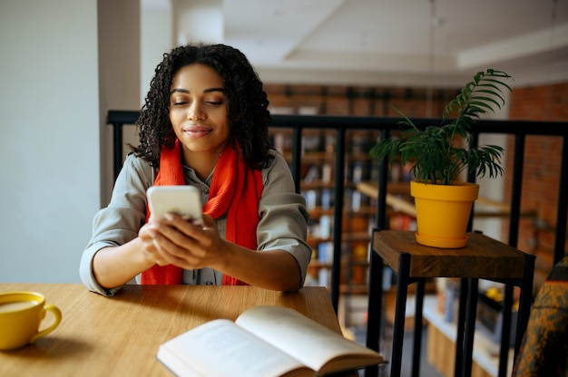 Jolie étudiante tient à l'aide du téléphone dans le café de la bibliothèque. Femme apprenant une matière, une éducation et des connaissances. Fille qui étudie dans la cafétéria du campus