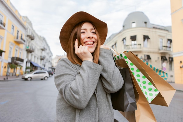 Jolie femme accro du shopping drôle en manteau d'automne et chapeau tenant de nombreux sacs à provisions en papier et parle au téléphone dans la ville européenne