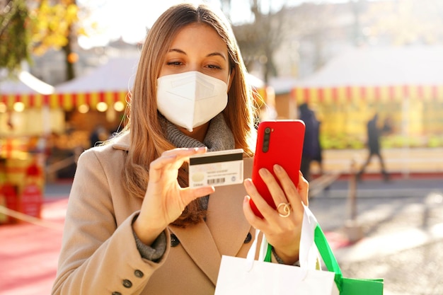 Jolie femme acheteuse portant un masque médical surveillant son numéro de carte de crédit pour faire des achats en ligne sur un téléphone portable avec un arrière-plan flou de Noël
