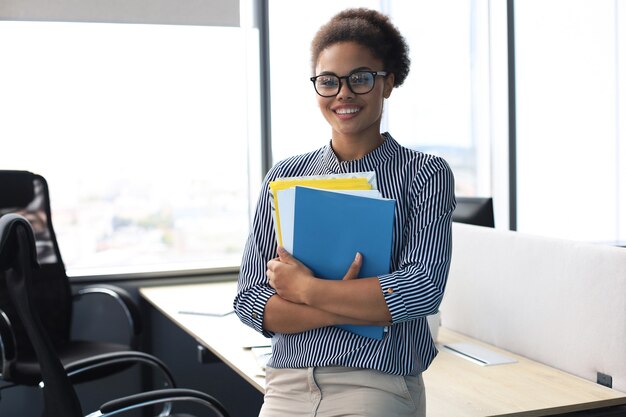 Jolie femme d'affaires afro-américaine regardant la caméra et souriante en se tenant debout au bureau.