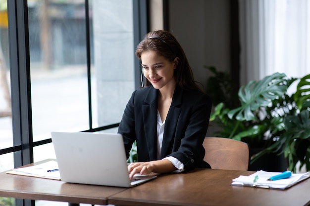 Jolie femme d'affaires asiatique en costume et casque souriant tout en travaillant avec un ordinateur