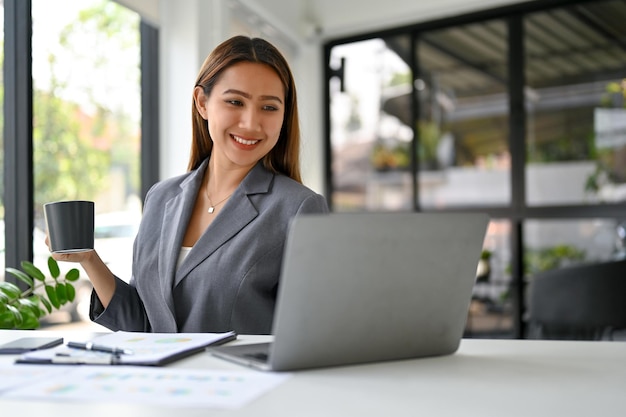 Jolie femme d'affaires asiatique en sirotant un café et travaillant sur ses tâches professionnelles sur un ordinateur portable