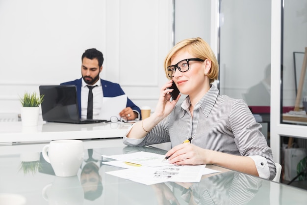 Jolie femme d'affaires blonde porte des lunettes parler par téléphone avant un homme barbu travaillant avec un ordinateur portable dans un intérieur de bureau blanc moderne