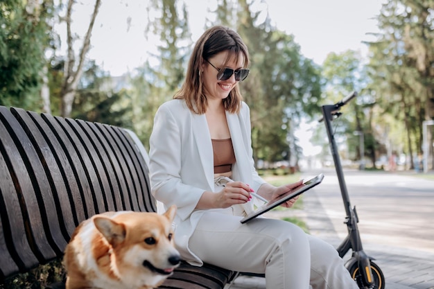 Jolie femme d'affaires en costume blanc assis dans un parc de la ville avec un chien Welsh Corgi Pembroke et travaillant sur une tablette numérique avec un scooter électrique sur fondxA