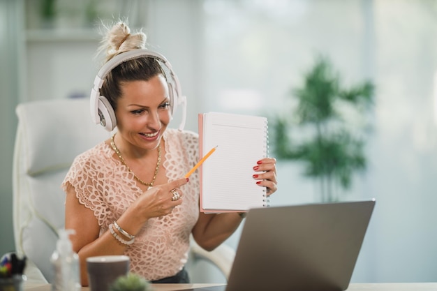 Une jolie femme d'affaires avec des écouteurs assise seule dans son bureau à domicile et travaillant en ligne sur son ordinateur portable.