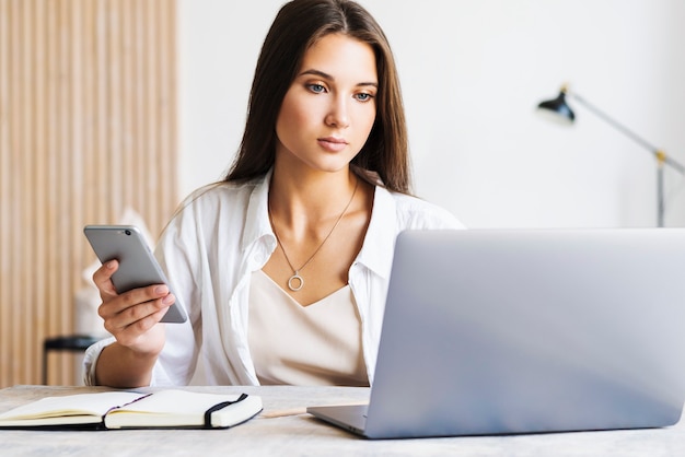 Photo jolie femme d'affaires est assise à table dans un ordinateur portable, le smartphone se trouve sur la table, prend des notes avec un crayon dans un ordinateur portable.