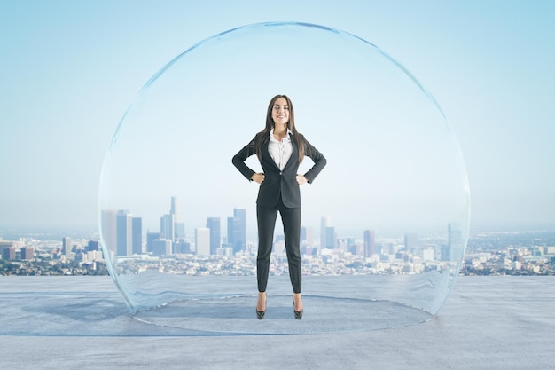 Jolie femme d'affaires européenne heureuse avec les bras croisés debout sous et à l'intérieur de la grande couverture de sphère de verre sur le sol en béton et le ciel lumineux fond de ville Concept de protection et de limitation