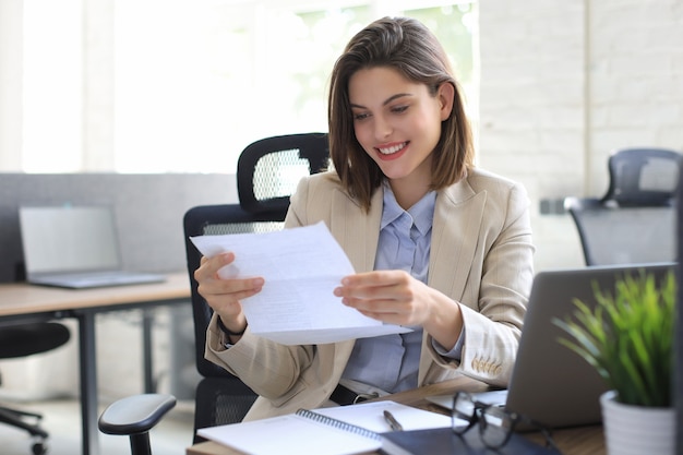 Jolie femme d'affaires joyeuse vérifiant les documents papier au bureau, travaillant sur un ordinateur portable.