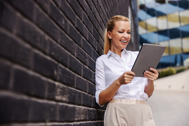 Jolie femme d'affaires à la mode blonde souriante innovante debout devant un mur noir et à l'aide de tablette.