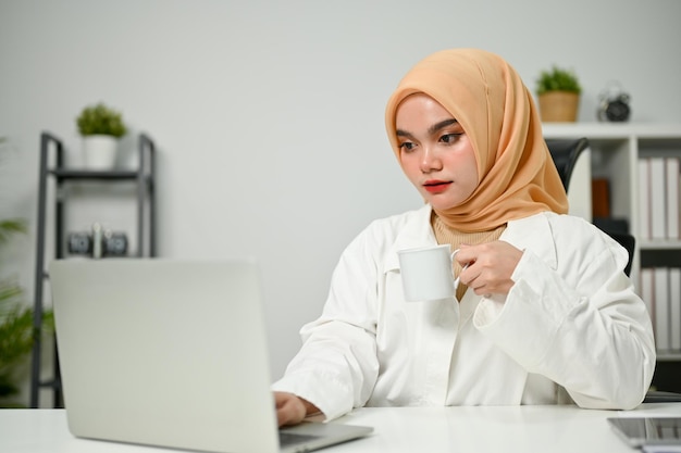 Jolie femme d'affaires musulmane asiatique regardant un écran d'ordinateur portable et sirotant un café