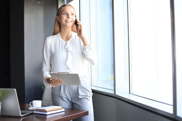 Jolie femme d'affaires parlant avec des collègues sur le téléphone portable en se tenant près de la fenêtre du bureau.