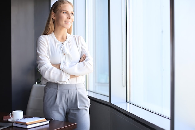 Jolie femme d'affaires regardant la caméra et souriante en se tenant debout dans le bureau près de la fenêtre.