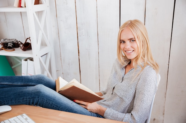 Jolie femme d'affaires souriante lisant un livre assis sur le lieu de travail au bureau