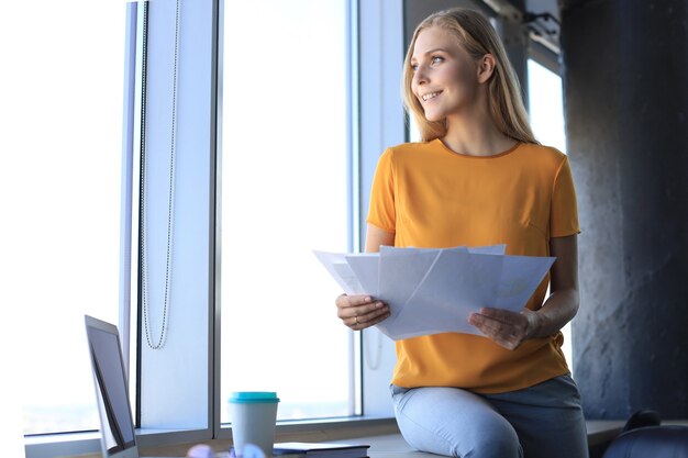 Jolie femme d'affaires tenant des documents et les regardant tout en étant assis sur le bureau au bureau.