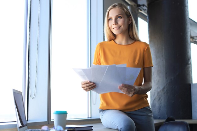 Jolie femme d'affaires tenant des documents et les regardant tout en étant assis sur le bureau au bureau.