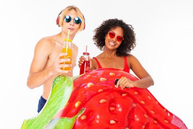 Jolie femme africaine et homme blond caucasien se tient en maillot de bain avec des matelas de plage en caoutchouc, boit du jus et des sourires, isolé sur fond blanc