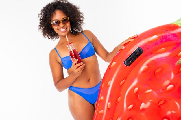 Jolie femme africaine se dresse en maillot de bain avec matelas de plage en caoutchouc fraise, écouter de la musique, boit du jus et des sourires isolé sur fond blanc