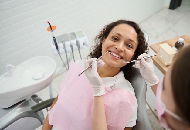 Jolie femme afro-américaine avec un beau sourire à pleines dents et des dents blanches saines souriantes regardant la caméra à côté d'un dentiste utilisant des outils dentaires stériles lors d'un examen planifié dans une clinique dentaire