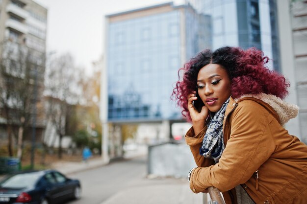 Jolie femme afro-américaine bouclée en manteau marron posée près des garde-corps contre un bâtiment moderne à plusieurs étages parlant au téléphone mobile