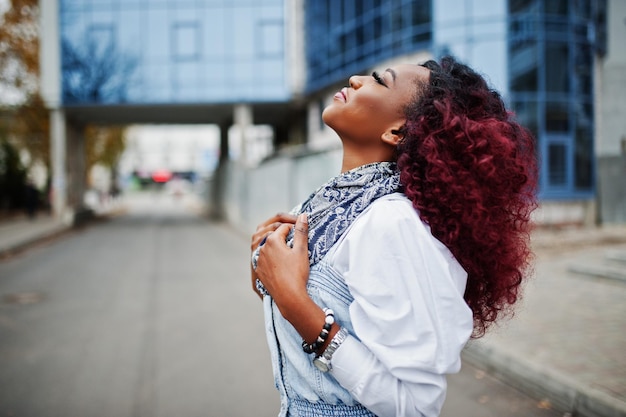 Jolie femme afro-américaine bouclée en robe jeans posée contre un bâtiment moderne à plusieurs étages