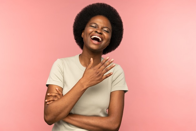 Jolie femme afro-américaine avec une coiffure afro riant d'une blague avec les yeux fermés sur un mur rose