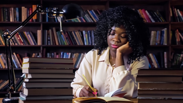 Une jolie femme afro-américaine avec une coiffure écrit dans un cahier en papier près d'une pile de livres et d'une lampe à table contre des étagères dans une pièce semi-obscure